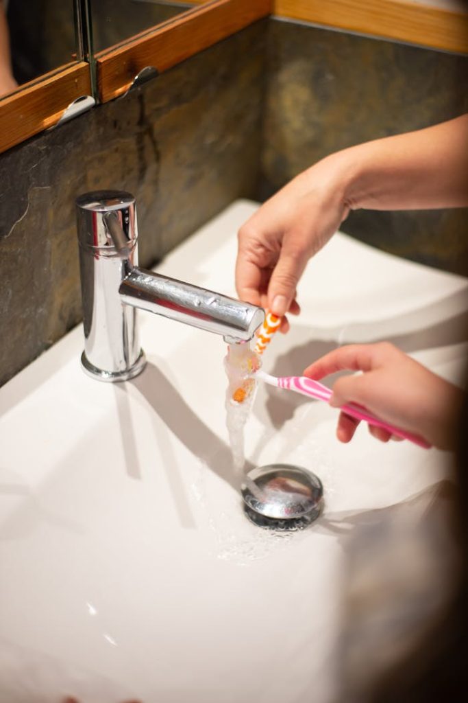 pexels photo 7492911 Close-up of hands washing toothbrushes under running faucet in a bathroom setting.